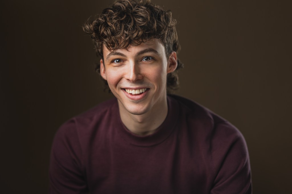 peter sarty headshot smiling into the camera on a dark brown backdrop. he is wearing a burgundy mock turtle neck. 