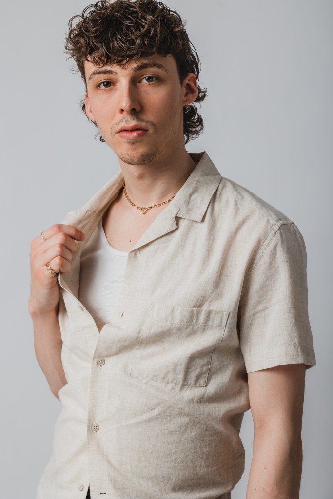 peter sarty headshot looking straight into the camera. he is wearing a linen button down dress shirt and small gold chain. 