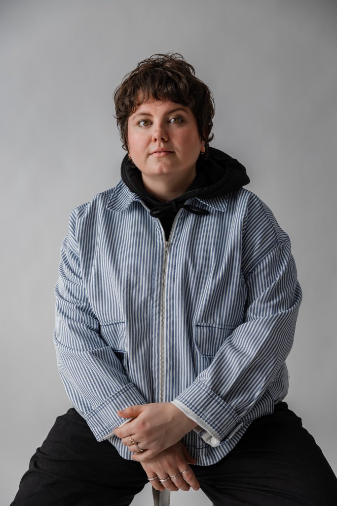 stevey wearing a striped blue jacket with their hands crossed in front of them. they look right into the camera on a white backdrop. 
