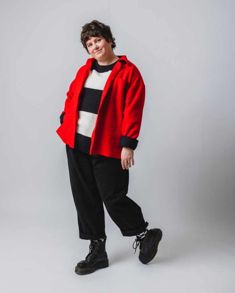stevey hunter full body shot wearing a striped white and black sweater underneath a bright red jacket on a white background. 