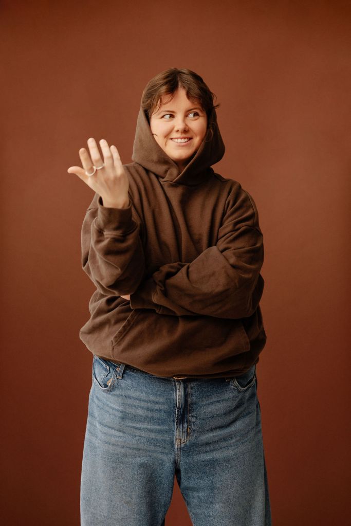 Stevey with a brown hoodie on with a cheeky smile looking off to the distance with their hand up in front of a brown background.