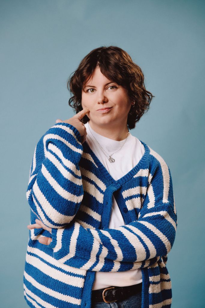 Stevey on a light blue background with their finger pointed cheekily into their cheek with a striped blue and white cardigan with one button done up.